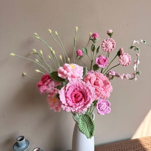 K&ouml;nnte beinhalten: Ein Strau&szlig; geh&auml;kelter rosa Blumen in einer wei&szlig;en Vase. Die Blumen sind aus Garn gefertigt und haben einen zarten, femininen Look. Die Vase ist schlicht und elegant, und die Blumen sind auf eine sch&ouml;ne, nat&uuml;rliche Weise angeordnet.