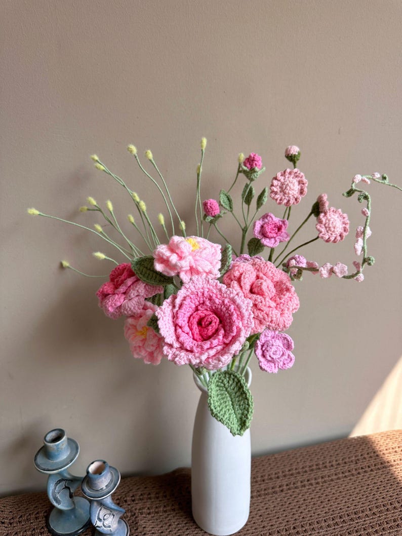 K&ouml;nnte beinhalten: Ein Strau&szlig; geh&auml;kelter rosa Blumen in einer wei&szlig;en Vase. Die Blumen sind aus Garn gefertigt und haben einen zarten, femininen Look. Die Vase ist schlicht und elegant, und die Blumen sind auf eine sch&ouml;ne, nat&uuml;rliche Weise angeordnet.