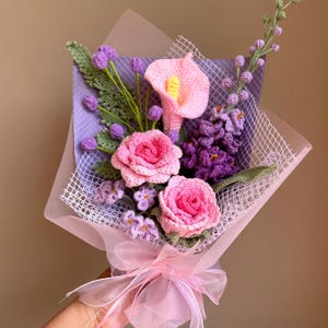 K&ouml;nnte beinhalten: Ein geh&auml;kelter Blumenstrau&szlig; in Rosa und Lila. Der Strau&szlig; enth&auml;lt Rosen, Calla-Lilien und kleinere lila Bl&uuml;ten. Die Blumen sind in ein rosa-wei&szlig;es Netz gewickelt.
