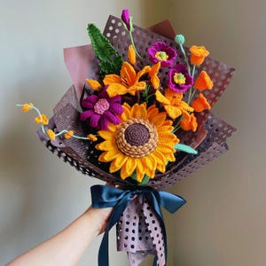 Può includere: Un bouquet all'uncinetto fatto a mano con un grande girasole giallo, gigli arancioni e fiori viola. Il bouquet è avvolto in carta marrone e legato con un nastro blu scuro. I fiori sono fatti di filato e hanno trame dettagliate.