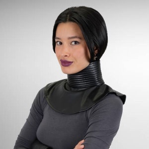 May include: A black leather choker with a ribbed texture. The choker is worn by a person with short black hair and dark lipstick.