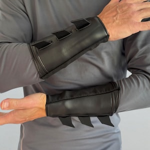 Könnte beinhalten: Ein Paar schwarze Unterarmschoner mit glatter, matter Oberfläche und drei markanten, dreieckigen, flossenartigen Vorsprüngen an der Außenkante jeder Manschette. Die Schoner zeichnen sich durch ein strukturiertes Design aus.
