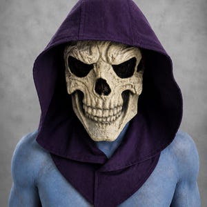 May include: A skull mask with deep eye sockets and a prominent jawline, worn under a dark purple fabric hood. The mask features an off-white, bone-like texture with realistic shading. The wearer has blue skin visible on the chest and shoulders.