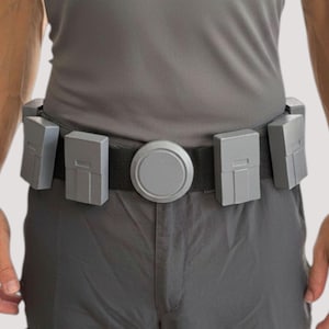 Op de afbeelding: Een zwarte utility-riem met een centrale ronde gesp en vier rechthoekige grijze tasjes bevestigd aan de band. De tasjes hebben een gladde, matte afwerking met subtiele verdiepte details aan de voorzijde en zijn bevestigd aan een zwarte stoffen band.