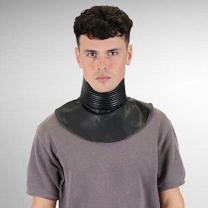 May include: A black faux leather neck gaiter featuring a high, ribbed, stand-up collar that transitions into a smooth, rounded bib-style base that covers the upper chest and shoulders, worn over a plain, short-sleeved, solid gray t-shirt.