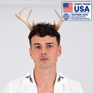 May include: A headband featuring two light brown, textured deer antlers. The wearer is dressed in a white collared shirt with small, colorful acorn and leaf illustrations embroidered on the collar points. The image includes a blue and white graphic.