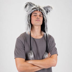 Puede incluir: Un gorro de animal de piel sintética con fibras largas y desgreñadas de color gris y blanco, orejas puntiagudas prominentes y dos largos cordones trenzados negros que cuelgan a los lados, terminados con pompones de piel sintética.