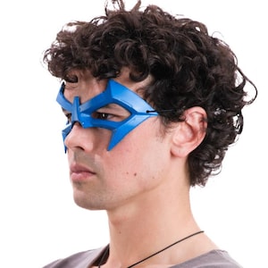 May include: A bright blue moulded eye mask with sharp, angular edges and a pointed design that covers the eye area. The mask features a thin black elastic strap that secures around the head, providing a snug fit across the bridge of the nose.