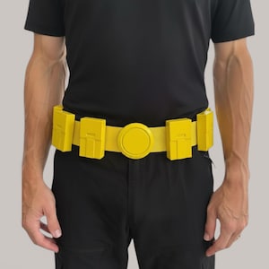 May include: A bright yellow utility belt featuring a central circular buckle and four rectangular pouches attached to the strap. The belt is worn around the waist over a black t-shirt and black trousers, showcasing a functional design with distinct compartments.