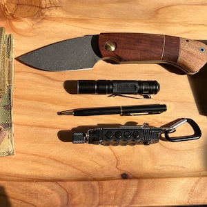 Puede incluir: Un cuchillo plegable con mango de madera, una linterna negra, un bolígrafo negro, una multiherramienta negra con mosquetón y un encendedor Zippo dorado en una caja negra sobre una superficie de madera.