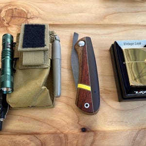 Puede incluir: Una linterna verde, una bolsa de cuero marrón con un parche negro, una navaja marrón y amarilla, y un encendedor dorado en una caja negra con el texto "Vintage Look" y "Zippo".