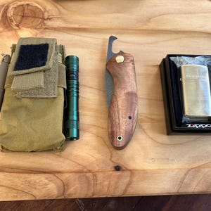 Könnte beinhalten: Eine Sammlung von Alltagsgegenständen, darunter eine beige Tasche mit Stift und grüner Taschenlampe, ein Messer mit Holzgriff und ein goldfarbenes Zippo-Feuerzeug in einer schwarzen Schachtel. Die Gegenstände sind auf einer Holzoberfläche angeordnet.