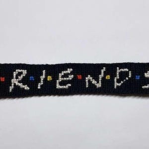 May include: Black friendship bracelet with the word "FRIENDS" woven in white thread. The bracelet has colorful dots in red, blue, yellow, and orange.