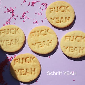 Könnte beinhalten: Fünf runde, hellgelbe Kekse mit den Worten "FUCK YEAH" eingeprägt, angeordnet auf einer lila Oberfläche. Rosa Zuckerkristalle sind um die Kekse herum verstreut. Der Text "Schrift YEAH" befindet sich unten.