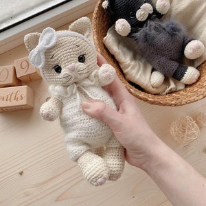 Peut inclure: Deux jouets en crochet en forme de chat. L'un est blanc avec un nœud rose et l'autre est noir avec un gilet en fourrure grise. Les deux ont un visage blanc avec des yeux noirs et un nez rose.