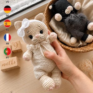 May include: Two crocheted cat dolls. One cat is cream-colored, wearing a white headband with a bow and a white ruffled collar. The second cat is black with a dark gray fuzzy belly, sitting in a woven basket lined with a light-colored cloth.