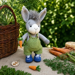 May include: A crocheted donkey stuffed animal with gray fur, a white muzzle, and a tuft of gray hair on its head. The donkey wears green knit overalls with small brown buttons and blue crocheted shoes, holding a small orange crocheted carrot.
