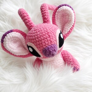 May include: A pink crochet plush toy featuring large ears with light pink centers and purple tips, two antennae, and a purple nose. The character has large black eyes with white highlights and a light pink chest patch, crafted from soft yarn.