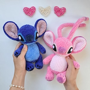 May include: Two plush crochet dolls, one dark blue and one pink, featuring large ears and round eyes. The blue doll has a lighter blue belly, while the pink doll has a lighter pink belly and long antennae. Three small woven hearts sit above.