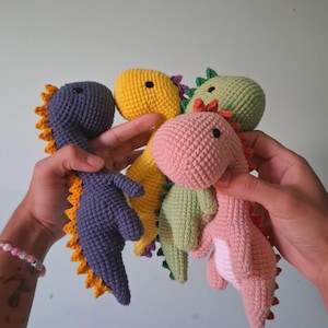 May include: A collection of four small crocheted dinosaur toys in various colours including dark grey with yellow spikes, bright yellow with purple accents, light green, and soft pink with a white belly and red spikes, all featuring black bead eyes.