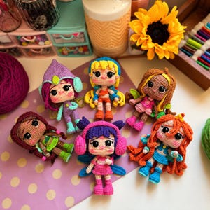 Könnte beinhalten: Sechs kleine Häkelpuppen mit großen schwarzen Augen und farbenfrohen Garnoutfits. Die Puppen haben verschiedene Haarfarben wie Lila, Gelb, Braun, Dunkelbraun, Blau und Orange sowie unterschiedliche Accessoires wie Hüte, Flügel und Ohrenschützer.