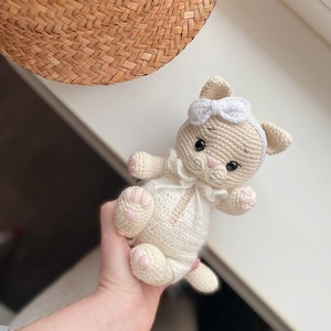 May include: A small, beige crocheted cat toy featuring a white ruffled collar and a white headband with a small bow. The cat has black embroidered eyes, a pink stitched nose, and light pink accents on its paws. The yarn texture is visible throughout.