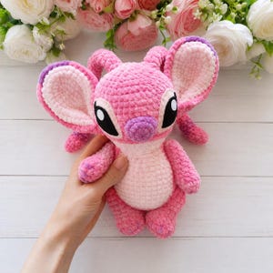 May include: A pink and light pink crocheted plush toy featuring large ears with purple accents, a rounded nose, and black and white eyes. The stuffed creature has a soft, textured yarn finish and is crafted with a detailed stitch pattern throughout.