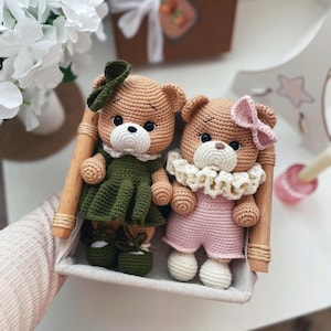 May include: Two handmade crochet teddy bears in a wooden tray. One bear wears a green dress and hat, the other a pink outfit and bow. Both have white collars and shoes. The bears are approximately 6 inches tall.