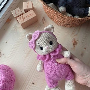 May include: A crocheted cat doll with cream-colored fur, wearing a bright pink romper with a ruffled collar and two small buttons, and a matching pink headband with a bow. The doll has embroidered facial features, including black eyes and whiskers.