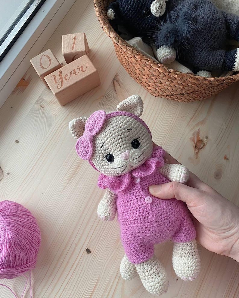 May include: A crocheted cat doll with cream-colored fur, wearing a bright pink romper with a ruffled collar and two small buttons, and a matching pink headband with a bow. The doll has embroidered facial features, including black eyes and whiskers.