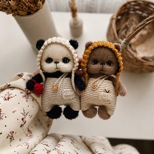May include: Two small, fuzzy, handmade plush animals resembling a panda and a brown bear. Both wear crocheted hoods and beige overalls with tiny bear-shaped buttons. The panda holds a small red heart, while the brown bear has a small pocket.