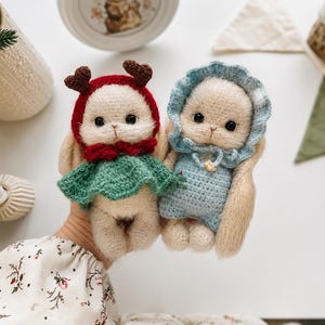 Könnte beinhalten: Zwei kleine, flauschige, cremefarbene Häkel-Hasenpuppen. Der linke Hase trägt eine rote Kapuze mit braunem Geweih und einen grünen Umhang. Der rechte Hase trägt eine hellblaue Haube und einen passenden hellblauen Strampler mit einer kleinen gelben Blume.
