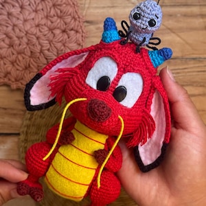 May include: A handcrafted crochet amigurumi figure of a red dragon with large ears, a yellow felt belly, and long yellow whiskers, featuring a small blue cricket figure perched on its head with black antennae and dark button eyes.