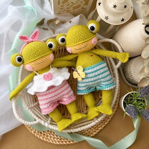 May include: Two crocheted frog dolls with large black eyes and pink cheeks. One frog wears a white ruffled top with a pink heart and pink striped shorts with a pink bow. The other frog wears blue and white striped shorts with a yellow butterfly.