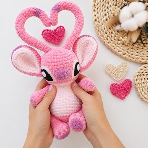 May include: A pink crocheted plush toy featuring large ears, black and white eyes, and two long antennae that curve to form a heart shape. The plush has a light pink belly and darker pink accents on the feet and ears, with a small pink woven heart nearby.