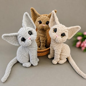 May include: Three plush crochet cat toys with large dark eyes and pink noses. One cat is light gray, one is brown, and one is beige. Each cat features pointed ears and a long tail, with the brown cat positioned inside a small woven basket.