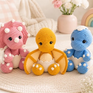 May include: Three handmade crochet dinosaur toys in pink, yellow, and blue. The pink triceratops, yellow pterodactyl, and blue dinosaur each feature white accents, black safety eyes, and are positioned sitting inside white cracked egg shells.