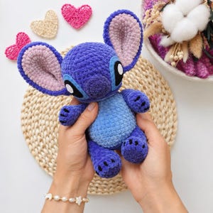 May include: A plush crochet toy of the character Stitch, featuring a dark purple body with a light blue belly, large ears with light pink inner sections, and black and white eyes. The soft, textured yarn construction gives the figure a fuzzy appearance.