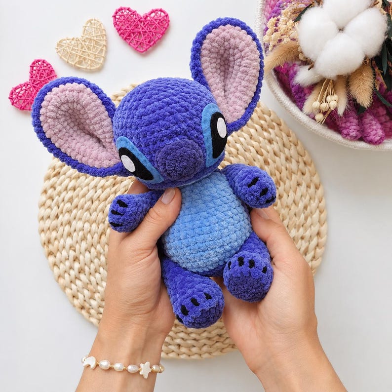 May include: A plush crochet toy of the character Stitch, featuring a dark purple body with a light blue belly, large ears with light pink inner sections, and black and white eyes. The soft, textured yarn construction gives the figure a fuzzy appearance.