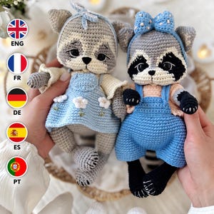 May include: Two handmade crochet raccoon dolls with large dark eyes and stitched facial features. The left raccoon wears a light blue dress with white flower embroidery and a matching headband. The right raccoon wears blue overalls and a blue polka-dot bow.
