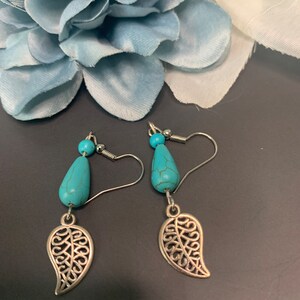 Puede incluir: Un par de pendientes turquesa en forma de lágrima con colgantes de hojas plateadas. Los pendientes tienen una pequeña cuenta turquesa y un gancho plateado. El fondo presenta una flor azul claro.