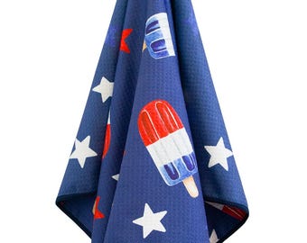 May include: A navy blue towel with a pattern of white stars and red, white, and blue popsicles. The towel is hanging from a white hook. The towel has a dark blue border.