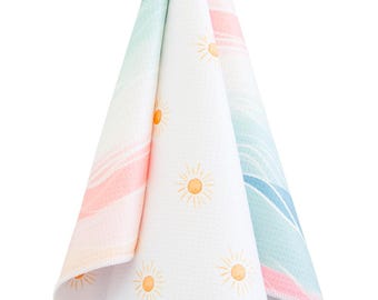 May include: A white cotton dish towel with a colorful ombre design of pink, blue, and green. The towel has a repeating pattern of small yellow suns.