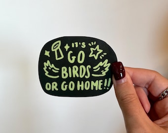 May include: A black sticker with the text "IT'S GO BIRDS OR GO HOME!!" in neon green. The sticker has a rounded shape and features illustrations of a football, stars, and wings. The sticker is held by a hand with a dark red nail polish.