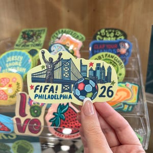 May include: A die-cut sticker featuring a silhouette of a boxer with arms raised, the Benjamin Franklin Bridge, the Philadelphia skyline, and a soccer ball. The text reads FIFA! PHILADELPHIA 26, accented with red and green stars on a cream background.
