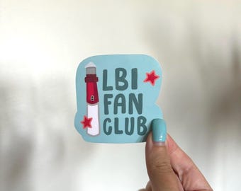 Può includere: Un adesivo con sfondo azzurro chiaro con un'illustrazione di un faro rosso e bianco. Il testo "LBI FAN CLUB" è in bianco e verde. Due stelle rosse sono sull'adesivo.
