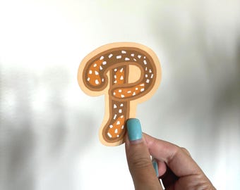 May include: A sticker in the shape of the letter "P" in brown, tan, and orange. The letter has a pretzel-like design with white speckles. The sticker is held by a hand with blue nail polish.