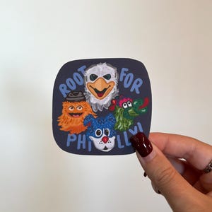 May include: A navy blue sticker with the words "Root for Philly" and illustrations of four cartoon characters. The characters include an eagle, a furry orange creature, a blue creature, and a green dragon-like creature.