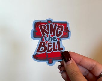 May include: A sticker in the shape of a bell with the words "Ring the Bell" in red and white. The bell is outlined in blue. The sticker is held by a hand with dark red nail polish.