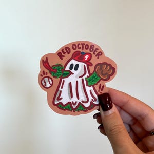 May include: A sticker featuring a cartoon ghost wearing a baseball cap and glove, with a baseball and green accents. The sticker has the words "Red October" in a circular design.
